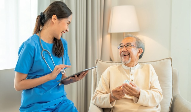 Nurse sits down next to elderly man while holding tablet using the MobiWork home & field health services software solution